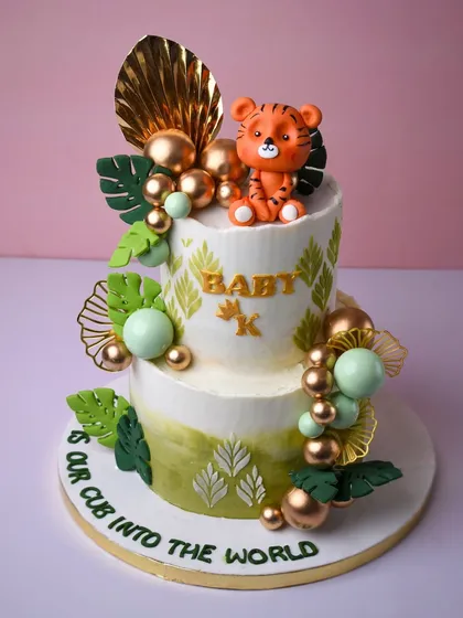 Welcoming a new cub into the world with this adorable tiger-themed baby shower cake. The two-tier design features jungle leaves, gold and green spheres, and a cute little fondant tiger sitting on top.