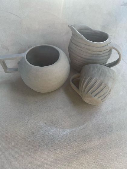 A set of unfired hand-built mugs and a pitcher. Each piece has a unique shape and texture, from the smooth, round mug to the carved and fluted smaller cup.
