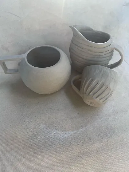 A set of unfired hand-built mugs and a pitcher. Each piece has a unique shape and texture, from the smooth, round mug to the carved and fluted smaller cup.