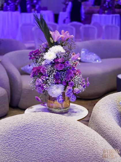 A beautiful floral arrangement of purple and white flowers in a gold vase, placed in a lounge area with plush, modern seating under purple lighting.