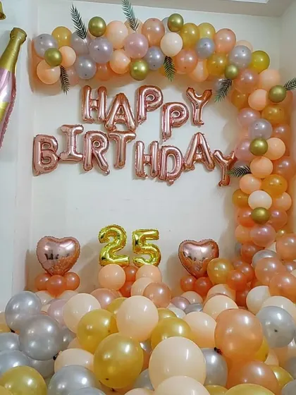 A 25th birthday home decoration with a balloon arch in peach, rose gold, and silver. The setup includes "Happy Birthday" and number foil balloons.