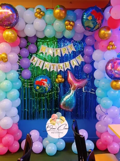 An under-the-sea mermaid theme birthday with a colorful balloon arch, shimmer curtain, and mermaid tail foil balloon.