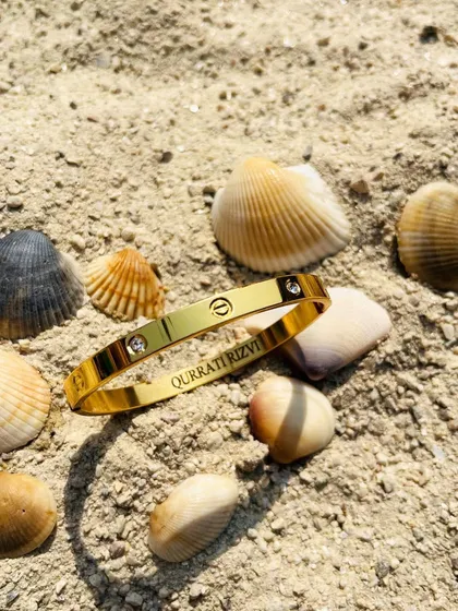 A personalized Cartier-style Kada in a gold finish, resting on the sand. The engraving inside adds a secret, personal touch to this classic design.