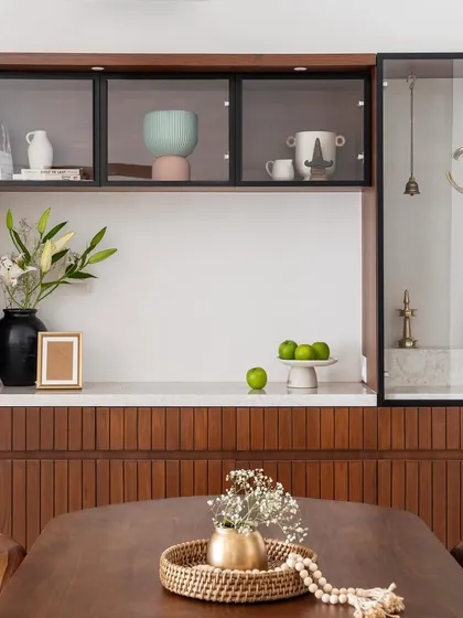 This custom unit serves as both a crockery cabinet and a puja space. The design features fluted wood paneling, a marble countertop, and glass-fronted upper cabinets.
