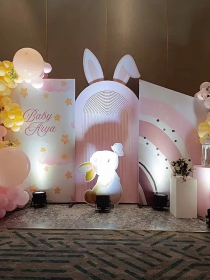 A full view of a bunny-themed stage for "Baby Arya." The design uses multiple backdrops, including a rainbow arch, and is accented with pink and yellow balloon garlands.