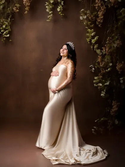 A regal portrait with a touch of sparkle. The mother-to-be wears a beautiful satin gown with gold glitter detail on her arm, set against a rustic floral backdrop.