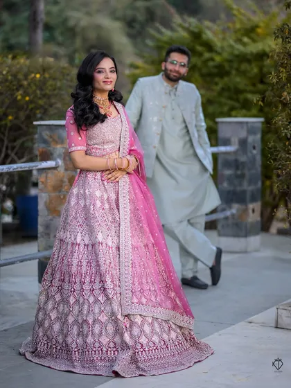 A creative portrait with the bride in the foreground, showcasing her beautiful lehenga, while the groom stands in the background.