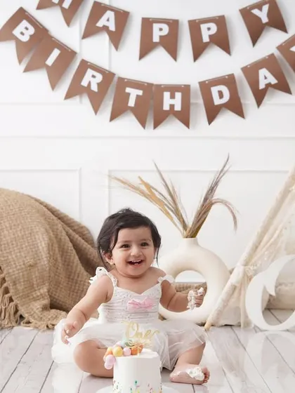 A happy baby girl enjoys her first birthday cake in a boho-themed setup, complete with a miniature tent and a 'One' sign.