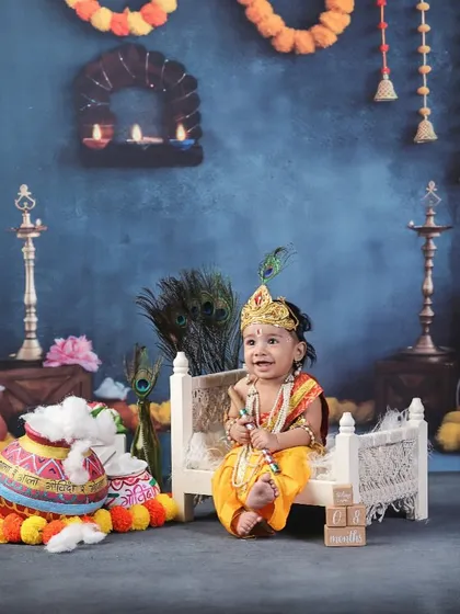 A wide shot of the elaborate Krishna setup, showing the baby seated on a small cot in a room decorated with marigolds, diyas, and idols.