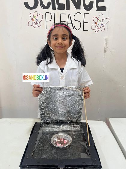 This young scientist is ready to cook with the sun! She carefully constructed her solar cooker using a box, aluminum foil, and plastic wrap to trap the sun's heat.
