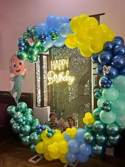 A Cocomelon themed birthday party with a circular balloon arch in shades of blue and yellow. A "Happy Birthday" neon sign adds a modern touch to the setup.