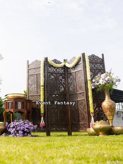 For an outdoor naming ceremony, I created this elegant and rustic setup. A carved wooden screen serves as a simple backdrop, accented with jasmine garlands and brass pots filled with purple flowers.