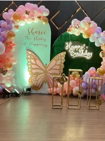 A spectacular butterfly themed first birthday celebration. This setup includes a large, glittery butterfly centerpiece, a light-up marquee number '1', and a beautiful balloon arch in pastel shades of purple, pink, and orange.