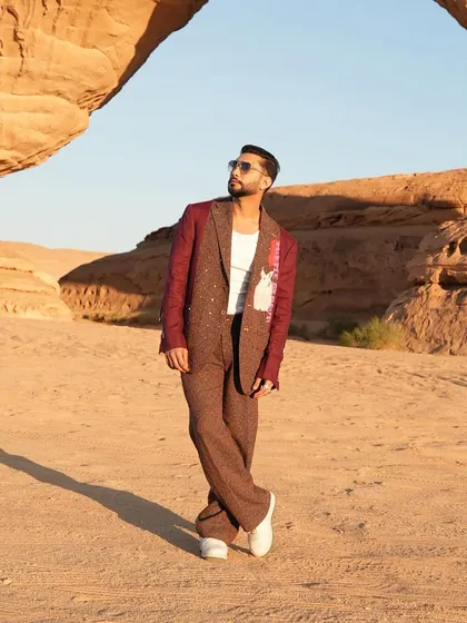 Zaid Darbar in a deconstructed suit against a desert landscape. The jacket features a unique combination of brown tweed with contrasting burgundy sleeves and graphic patches, creating a look that is both artistic and modern.
