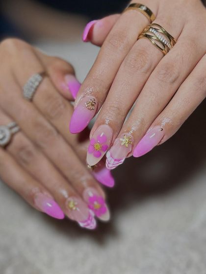 A vibrant pink ombre set on long stiletto nails, featuring 3D flowers, gold charms, and a marble accent nail.