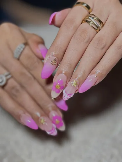 A vibrant pink ombre set on long stiletto nails, featuring 3D flowers, gold charms, and a marble accent nail.