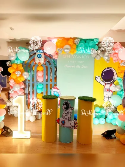 A fun and colorful space-themed first birthday stage for Shivank. The setup includes astronaut and rocket cutouts, planet-like balloon arrangements, and a "1" marquee light.