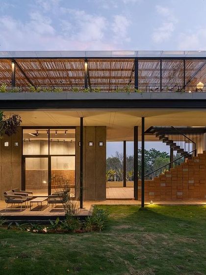 An outdoor seating area nestled under the building's large overhang, providing a shaded space to enjoy the landscape. The design seamlessly integrates indoor and outdoor living, with direct access to the lawn and views across the property.