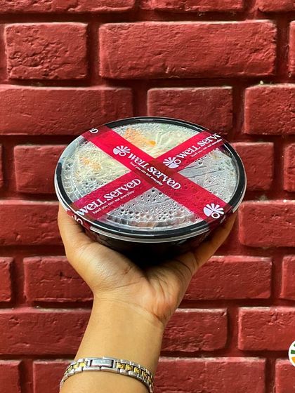 A hand holding a freshly packed meal bowl against a brick wall. This is my promise of delivering home-style meals straight to your office or home, every day.