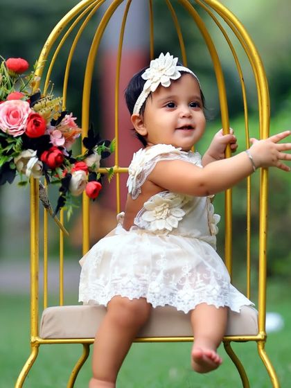 A lovely outdoor portrait of a baby girl in a white dress, sitting in a golden birdcage prop. The natural setting adds a whimsical touch.