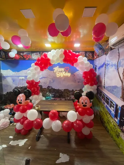 A Mickey Mouse themed birthday decoration in a restaurant with a red and white balloon arch and character sculptures.