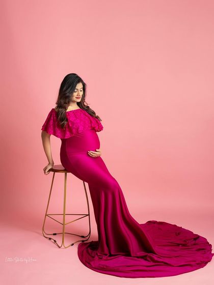 A vibrant and stylish portrait against a solid pink background. The mother-to-be is seated, wearing a sleek, form-fitting magenta gown with a cape-like shoulder detail and a long train.