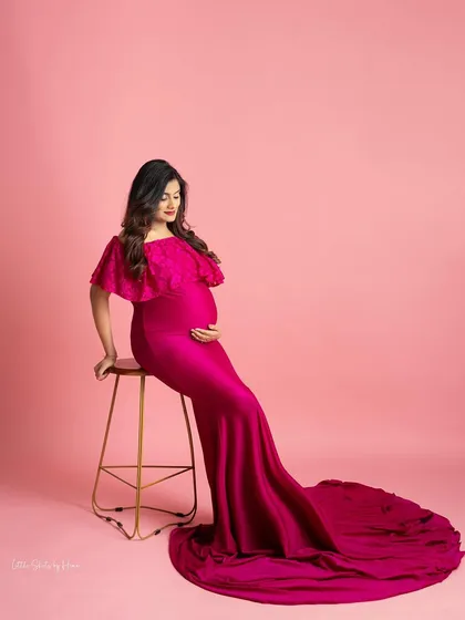 A vibrant and stylish portrait against a solid pink background. The mother-to-be is seated, wearing a sleek, form-fitting magenta gown with a cape-like shoulder detail and a long train.