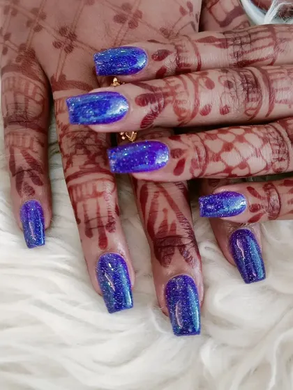 A dazzling set of square-shaped nails in a sparkling royal blue glitter polish, shown on henna-adorned hands.