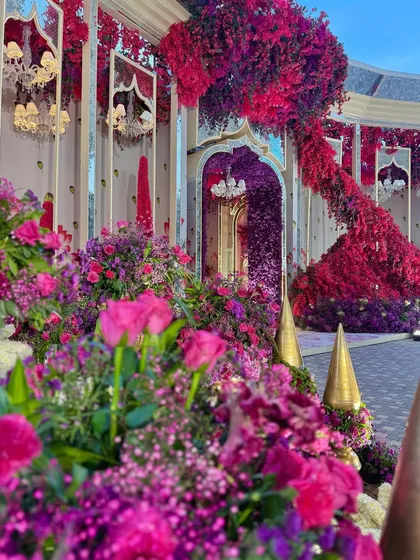 A view of the entrance, where a cascade of red flowers flows down the side of a mirrored arch, surrounded by a garden of purple blooms.
