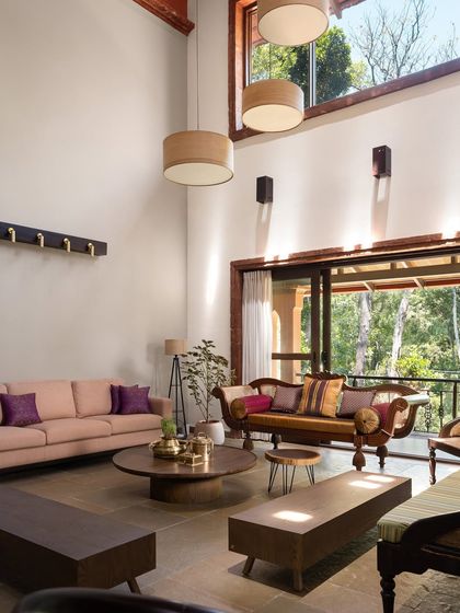 In this living area, a soaring twenty-foot ceiling is balanced by a warm, inviting furniture arrangement. A mix of modern sofas and a traditional carved rosewood settee creates a dialogue between old and new, all bathed in natural light from garden-facing doors.