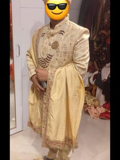 A client trying on a cream-colored sherwani with a matching stole and pearl necklace. Our team ensures a perfect fit and helps you accessorize for the complete look.