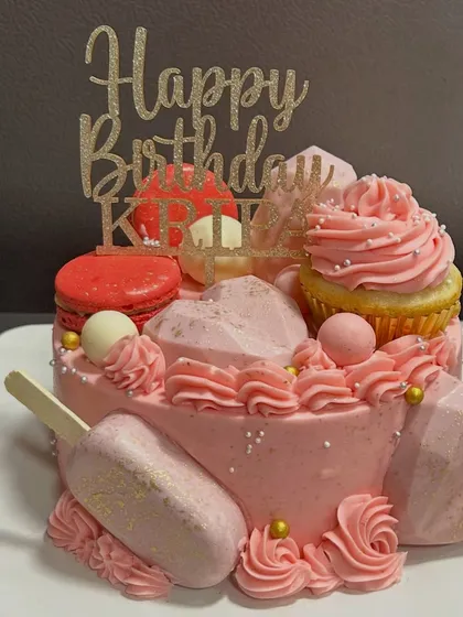 A fun and funky pink cake loaded with treats! It's topped with a cupcake, macarons, and geometric chocolate hearts for a truly indulgent birthday celebration.