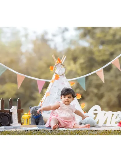 A beautiful setup for baby Anwita's first birthday photoshoot. The scene is complete with a teepee, bunting, a wooden fence, and a "One" sign, all arranged in a lovely park setting.