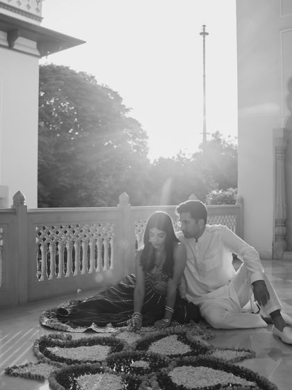 A quiet, tender moment captured in black and white. The couple sits on the floor of a palace veranda, their hands creating a floral rangoli, a beautiful symbol of their union.
