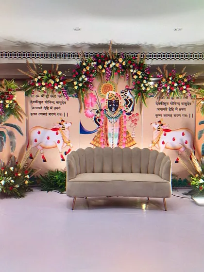 A beautiful stage designed for a religious ceremony, featuring traditional Pichwai art of Shrinathji and his cows. The decor is complemented by floral arrangements and banana leaf motifs, creating a serene and spiritual ambiance.