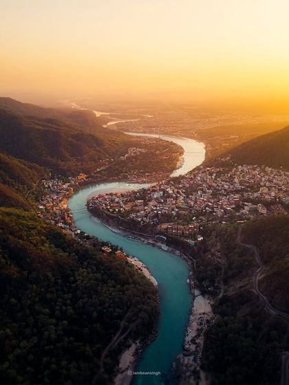 An epic drone shot of Rishikesh at sunset. The entire valley is bathed in a warm, golden light, with the Ganga river flowing like a ribbon of life through the landscape.