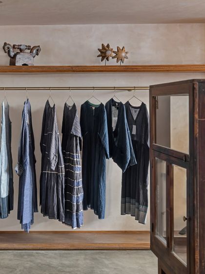 Clothing is displayed on simple wooden shelves with brass hanging rods, set against textured, lime-washed walls. The material palette is intentionally timeless, using natural wood and concrete to feel both refined and warm.