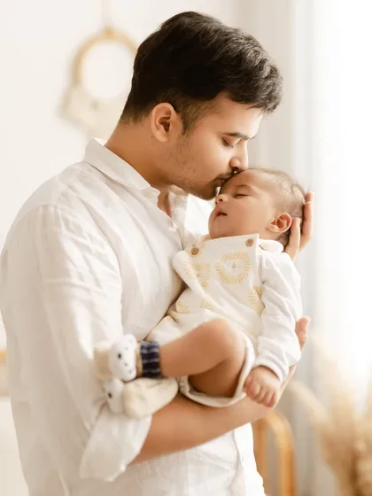 A father's gentle love shown in a simple gesture. He holds his sleeping baby and places a soft kiss on his forehead in this light-filled portrait.
