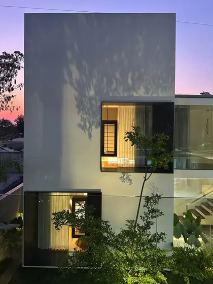 The house transforms from day to night. As evening approaches, the interior lighting creates a warm glow, and shadows from the trees dance across the minimalist white facade, adding a layer of natural art.