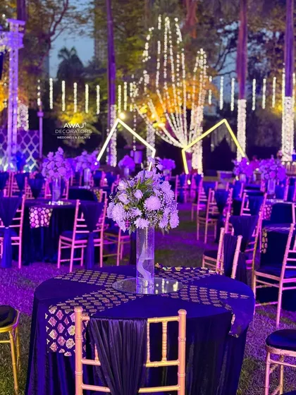 Elegant guest seating for a Sangeet, with deep blue tablecloths, gold-patterned runners, and simple floral centerpieces under a canopy of lights.