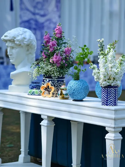 A modern-meets-classic vignette with a classical bust, set against a blue and white backdrop, and surrounded by colorful flowers in unique vases.