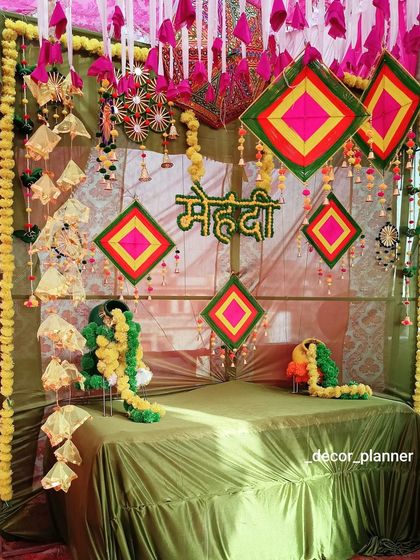 A beautiful Mehndi seating setup with an olive green theme. The backdrop is decorated with colorful handmade hangings and a 'Mehndi' sign, creating a lovely spot for the bride.