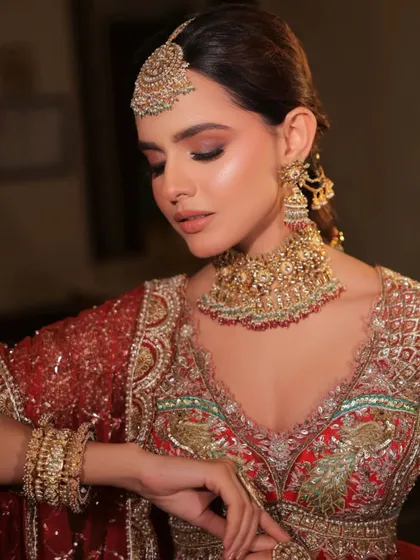 A soft, glamorous bridal portrait. The styling emphasizes the heavy gold choker and matching jhumkas, which perfectly complement the red and gold peacock embroidery on the blouse.