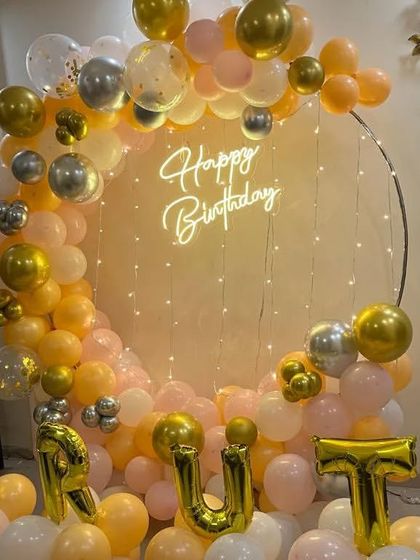 A warm and inviting balloon ring in peach, orange, and gold tones. This home setup includes a neon sign, fairy lights, and the name 'SHRUTI' spelled out in foil balloons on the floor.