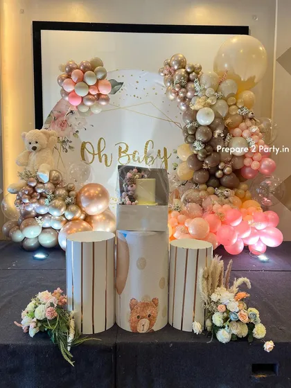 An elegant and modern "Oh Baby" themed baby shower backdrop. The setup features a sophisticated palette of rose gold, pink, and beige balloons, a plush teddy bear, custom pedestals for the cake, and delicate floral arrangements.