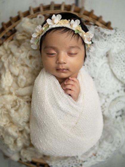 A dreamy portrait of a baby swaddled in white, nestled in a basket with soft, creamy textures. The floral headband adds a touch of nature.