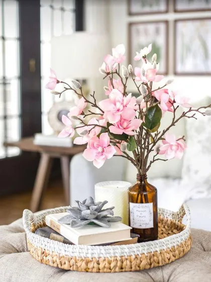 We styled this tabletop with a woven tray holding a collection of decorative items. A book, a succulent-shaped object, a candle, and a vase of artificial cherry blossoms come together to create a soft and elegant centerpiece.