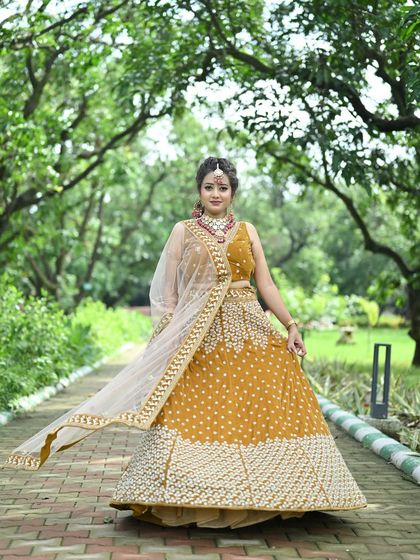 A model posing in the mustard yellow lehenga, her arms outstretched to show the full width and design of the outfit.