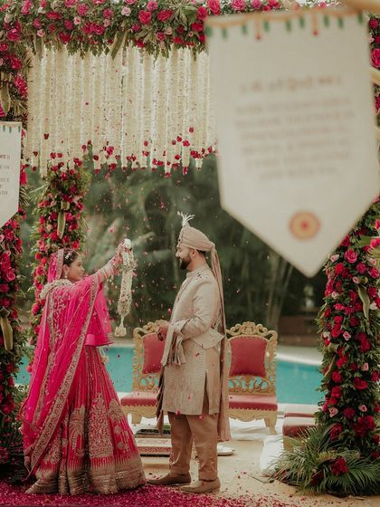 A wide shot of the playful Varmala exchange. This image shows the bride tossing petals at her groom, a fun and candid moment set against the beautiful poolside mandap.