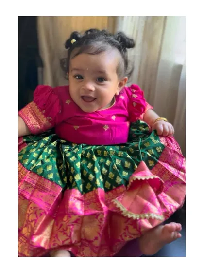 Even the tiniest members of the family can join in the festivities. This adorable baby girl is wearing a custom pattu pavadai in a classic green and pink combination, designed to be comfortable and beautiful.
